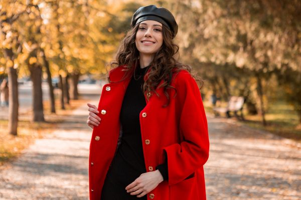 attractive stylish smiling woman with curly hair walking in park dressed in warm red coat autumn trendy fashion, street style, wearing beret hat